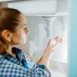 Female plumber in uniform installing drain pipe in the kitchen. Handywoman with toolbag repair sink, sanitary equipment service at home